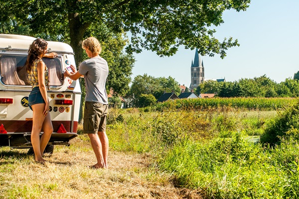 Twee mensen bij een retro caravan aan de rand van een veld, met uitzicht op het witte stadje Thorn en een kerktoren onder een heldere zomerlucht.