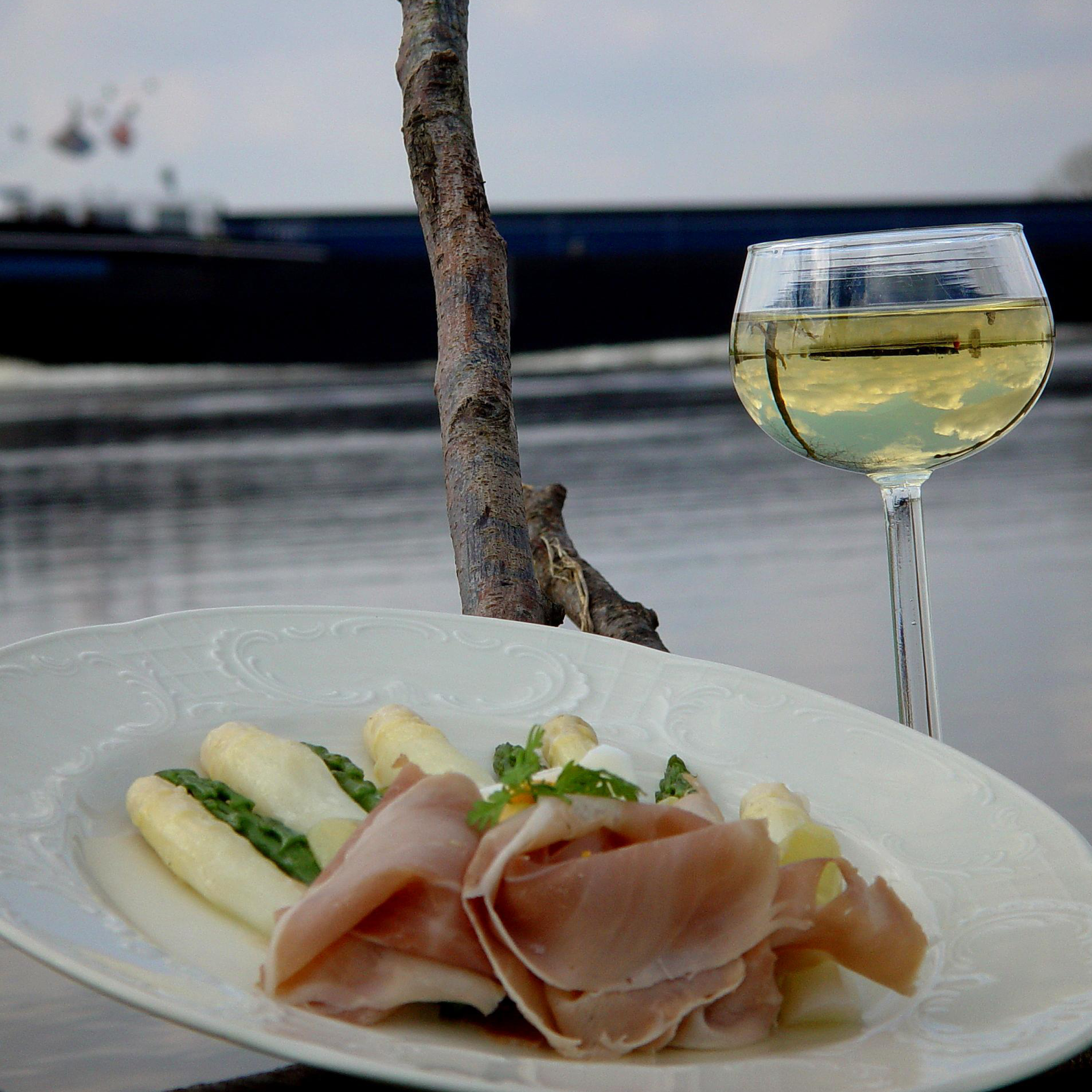 Bord met witte asperges en plakjes ham, geserveerd aan het water, met een glas witte wijn ernaast.