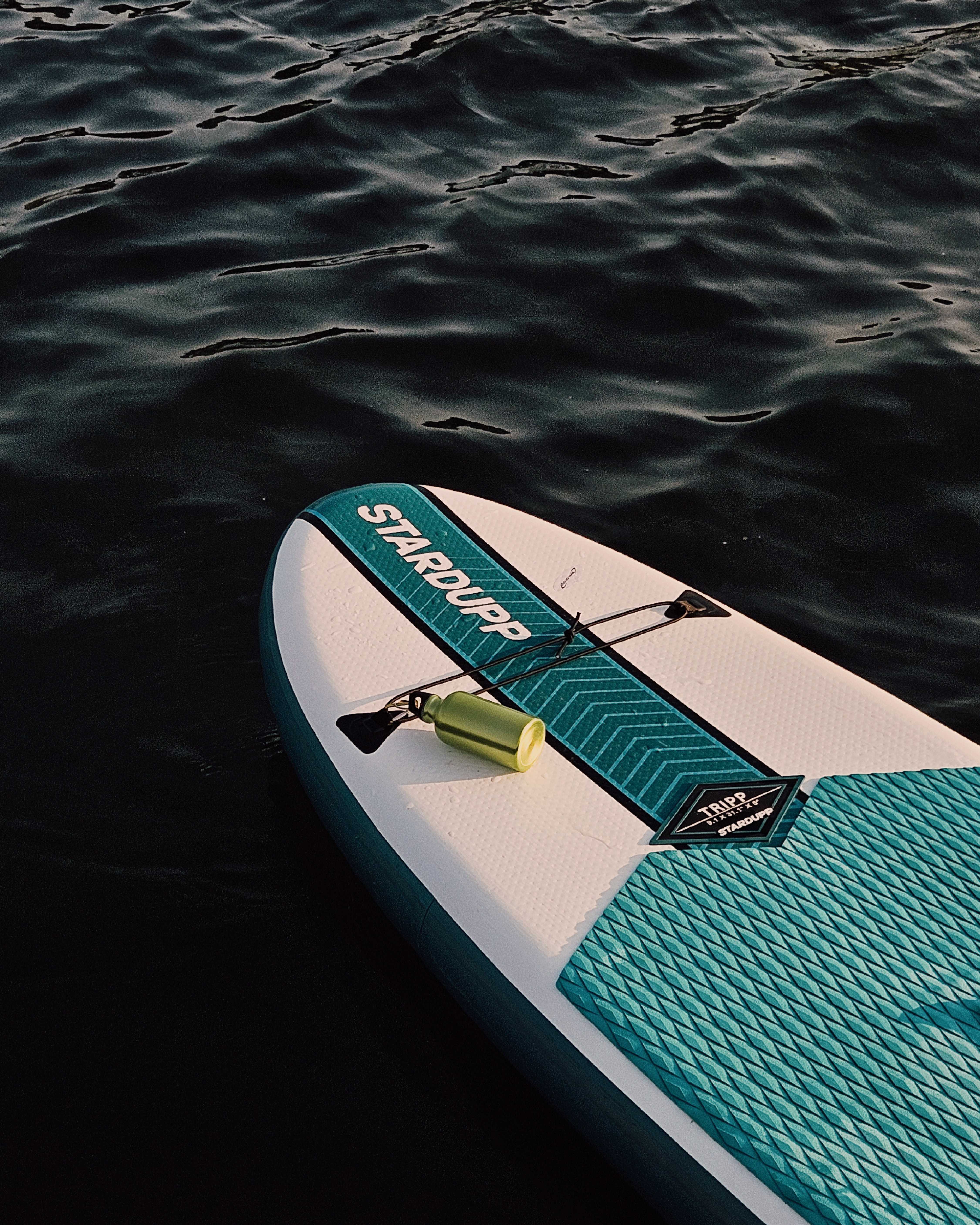 Opblaasbaar SUP-board van het merk Stardupp drijft op donker, kabbelend water, met een groene drinkfles vastgemaakt aan het elastiek aan de voorkant van het board.