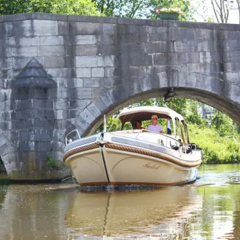 Sloep vaart onder een oude stenen brug op de Maasplassen, met mensen aan boord die genieten van een ontspannen vaartocht.