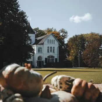 Wit landhuis op Landgoed Frymerson, omringd door hoge bomen en een groot grasveld, met pompoenen op de voorgrond in herfstlicht.