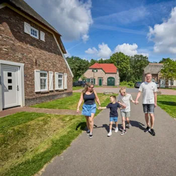 Gezin wandelt samen over een rustig pad in een groen bungalowpark met vrijstaande vakantiewoningen en blauwe lucht.