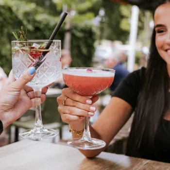 Twee personen proosten met cocktails op een terras, met een ontspannen sfeer en groen op de achtergrond.